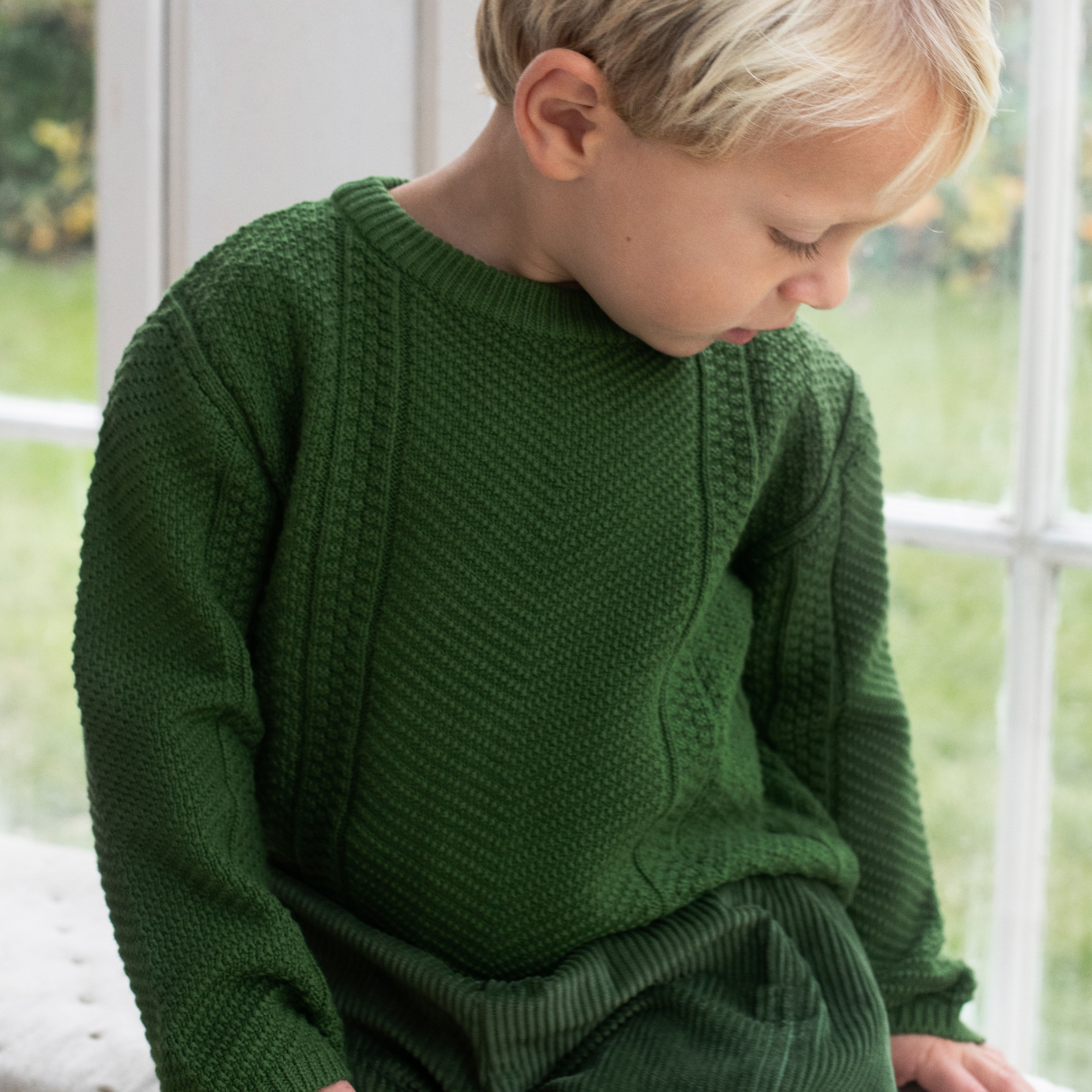 A young child with blonde hair wearing a dark green textured knit sweater and matching pants, sitting in front of a bright window.