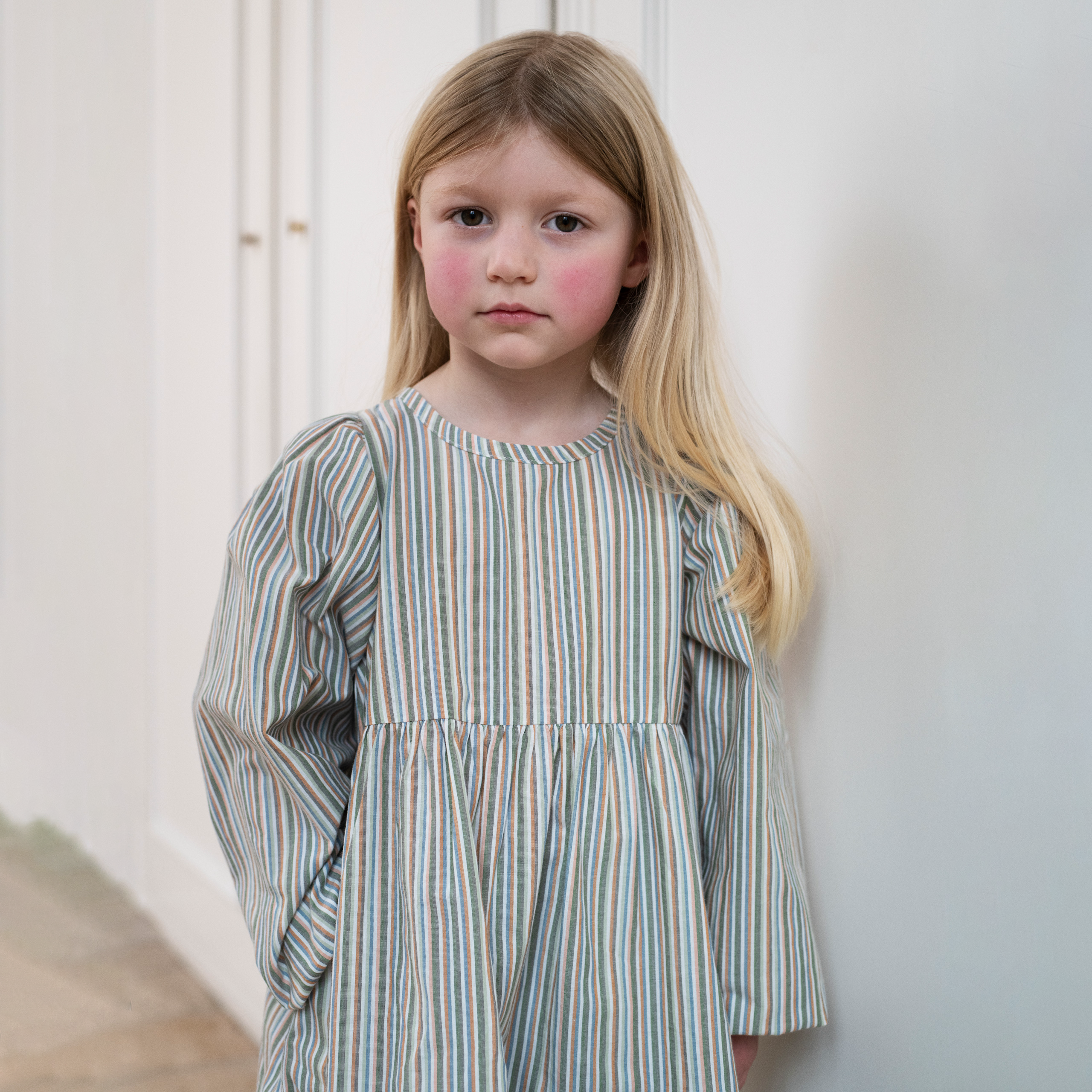 A young girl with long blonde hair stands against a white wall wearing a long-sleeved dress with vertical stripes in muted blue, green, orange, and cream.