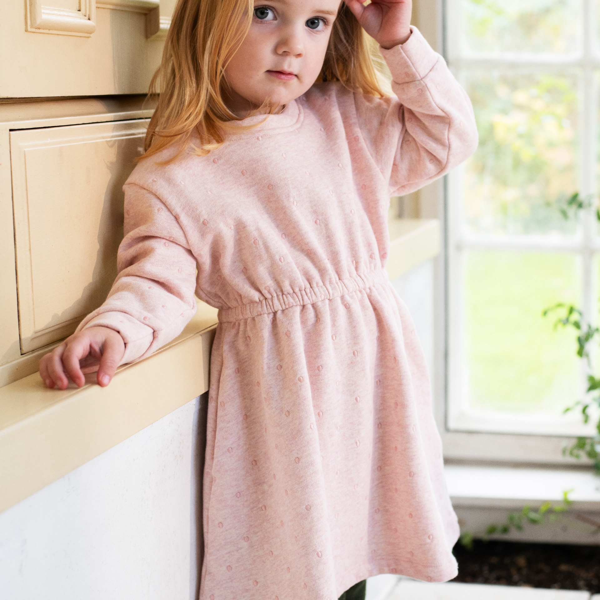 A young girl with blonde hair wearing a light pink long-sleeve dress with a gathered waist, leaning against a cream-colored wooden counter in a bright room.