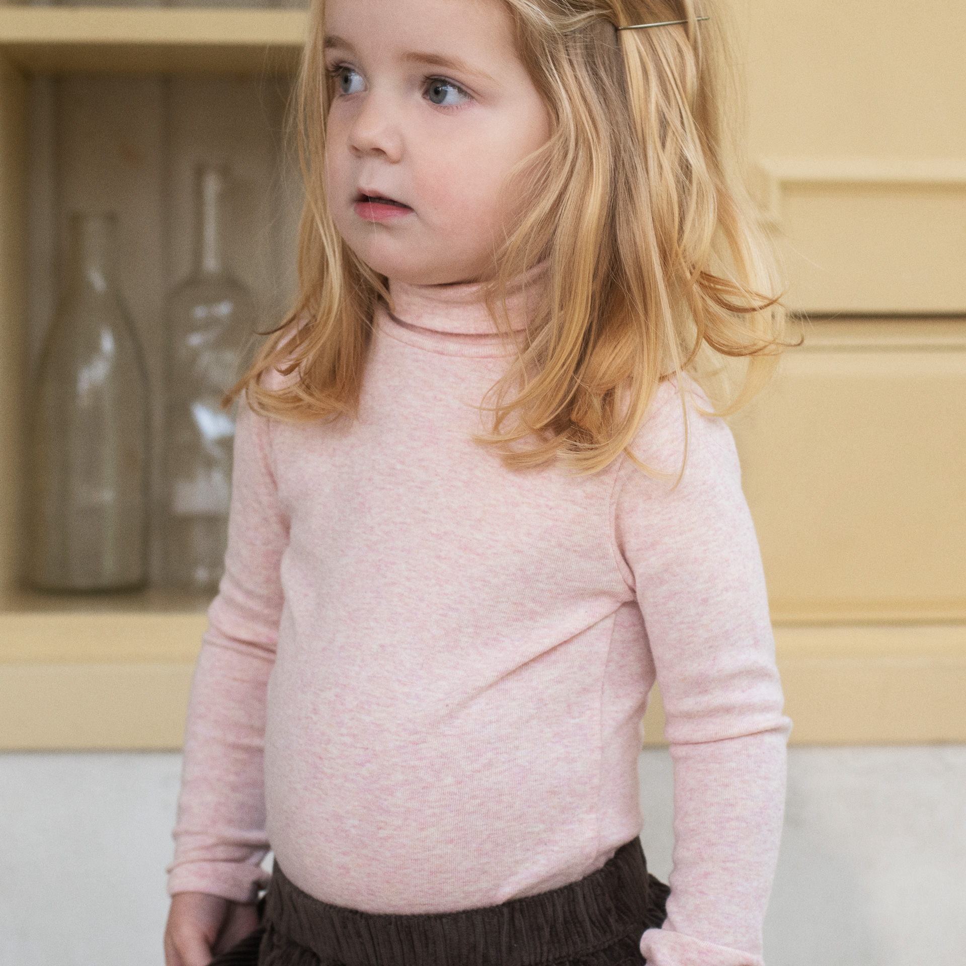 A young girl with blonde hair wearing a light pink long-sleeve turtleneck shirt, standing in front of a soft beige background.