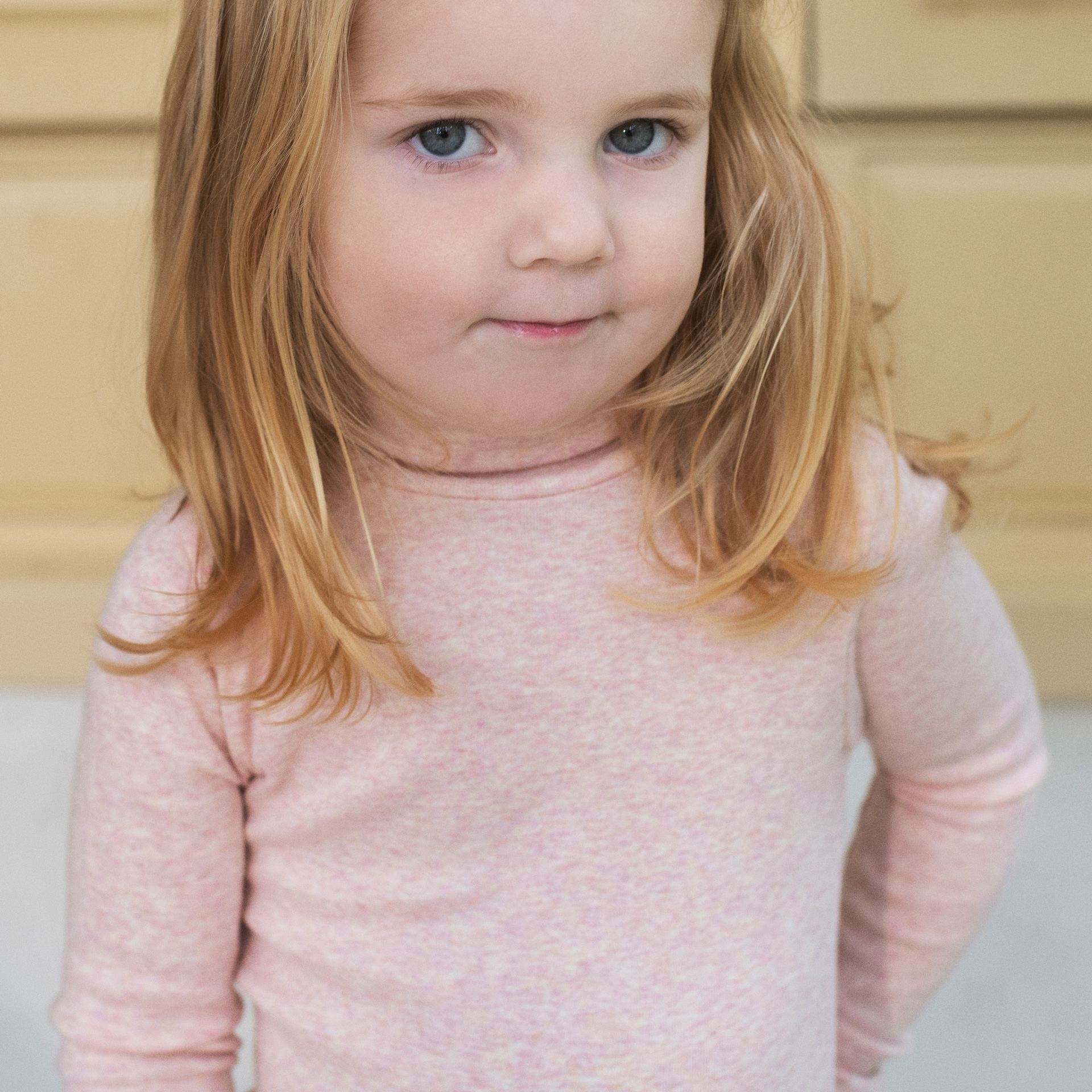 A young girl with blonde hair and blue eyes wearing a light pink long-sleeve turtleneck top, photographed in a warm indoor setting.
