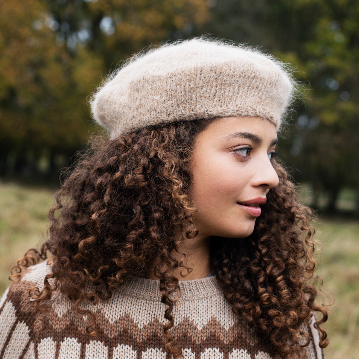 A woman with long, curly brown hair wearing a beige knit beret and a brown and beige patterned knit sweater, photographed outdoors against a blurred background of trees and grass.