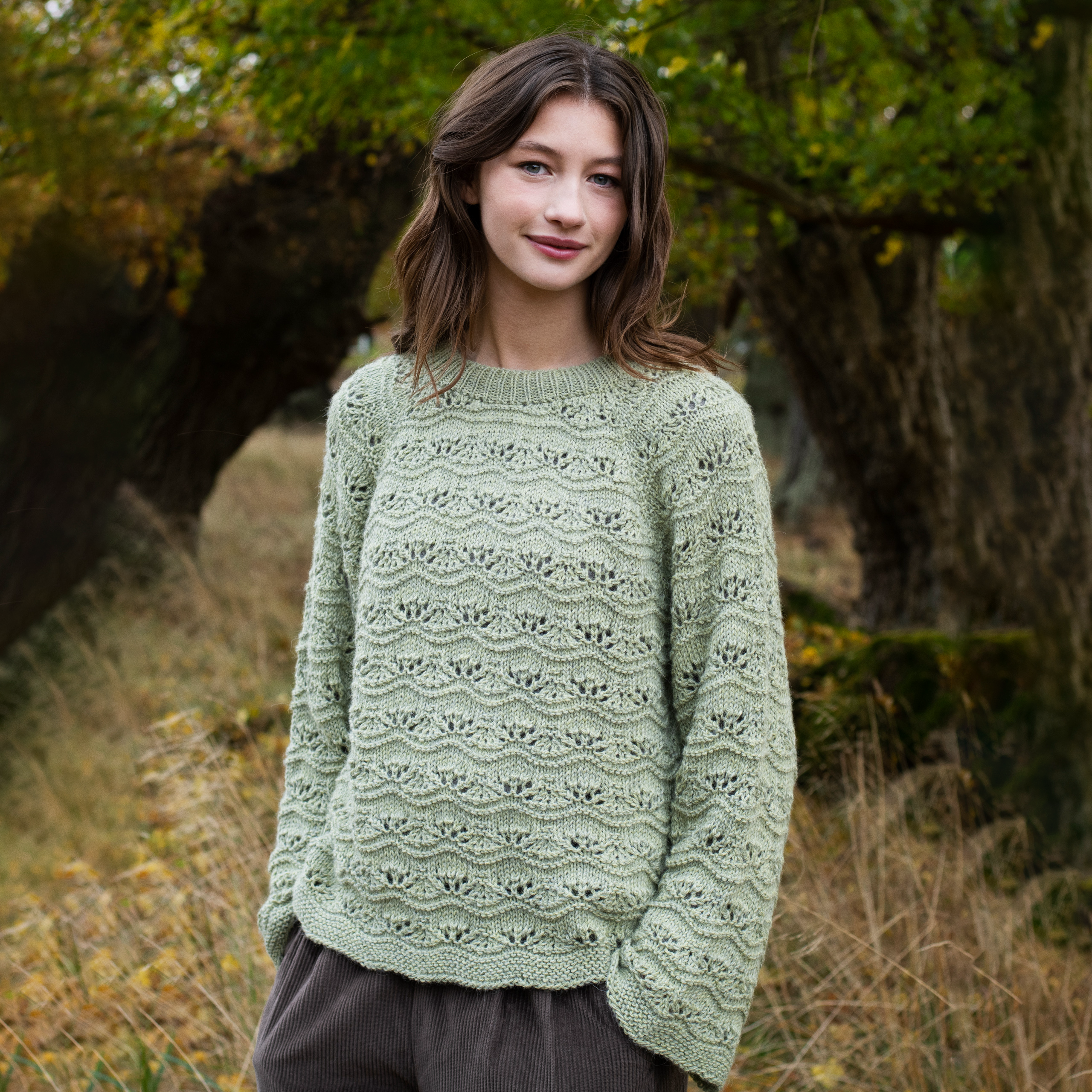 A young woman wearing a sage green, long-sleeve knit sweater with a textured openwork pattern, standing outdoors in a grassy, wooded area.