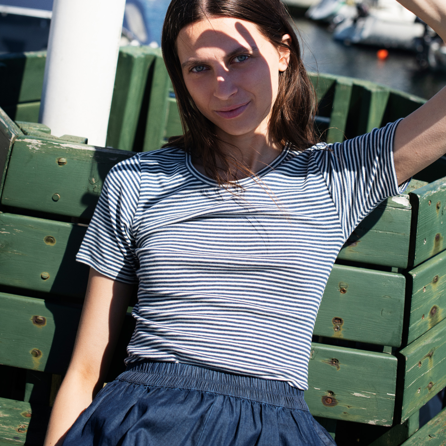 Woman in a striped shirt and blue skirt sitting on green wooden crates by a waterfront.