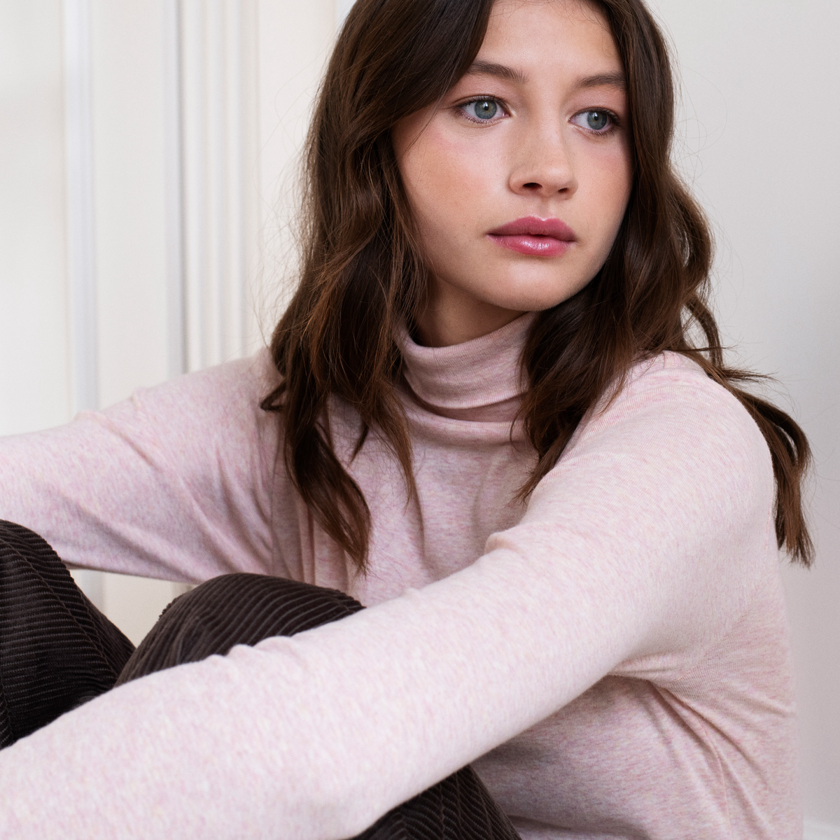A woman with long brown hair wearing a pale pink turtleneck and dark brown corduroy pants, sitting against a white paneled wall.