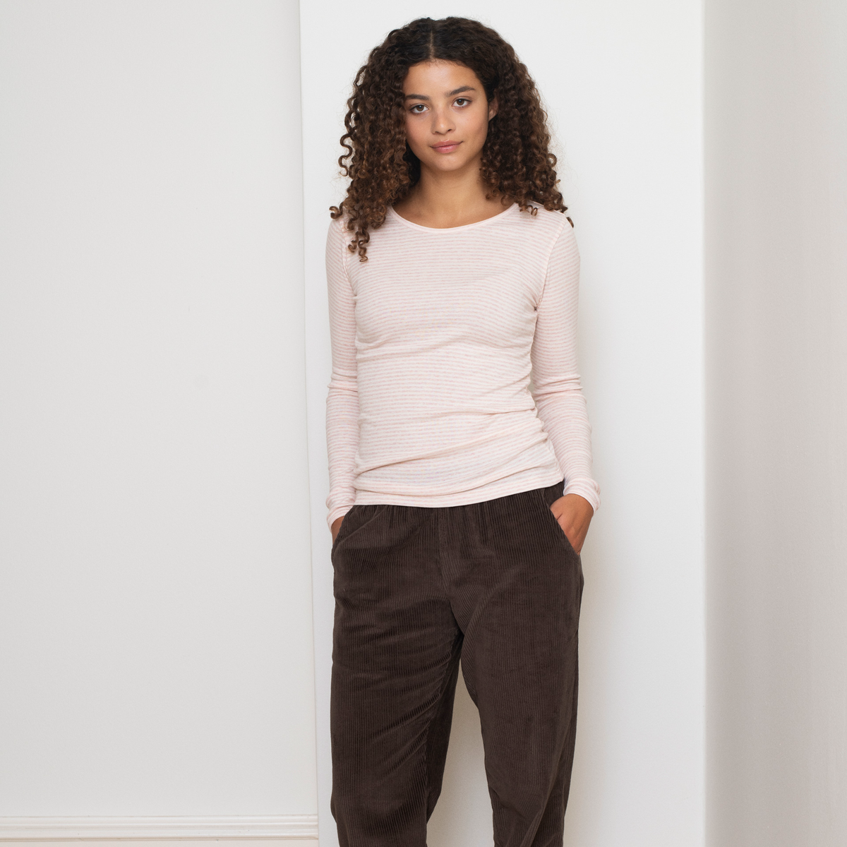 A woman with curly brown hair stands against a white wall wearing a pale pink ribbed long-sleeve top and dark brown corduroy pants.