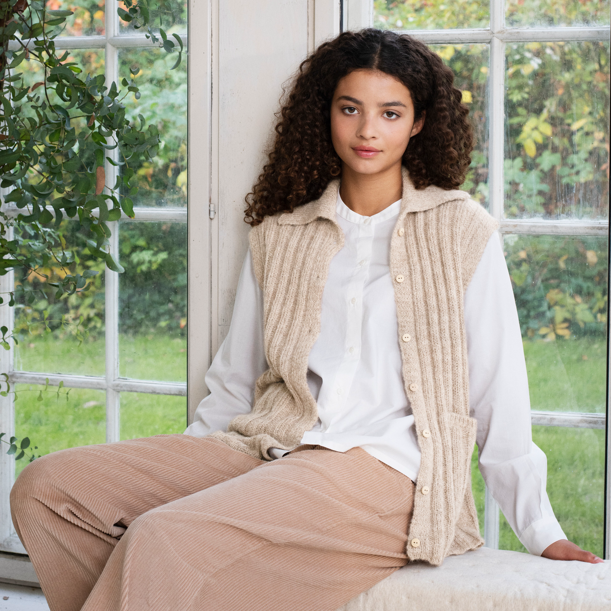 A woman with curly dark hair sits by a large white-framed window, wearing a beige ribbed knit sleeveless vest over a white button-up shirt and beige corduroy trousers. Greenery is visible outside the window.