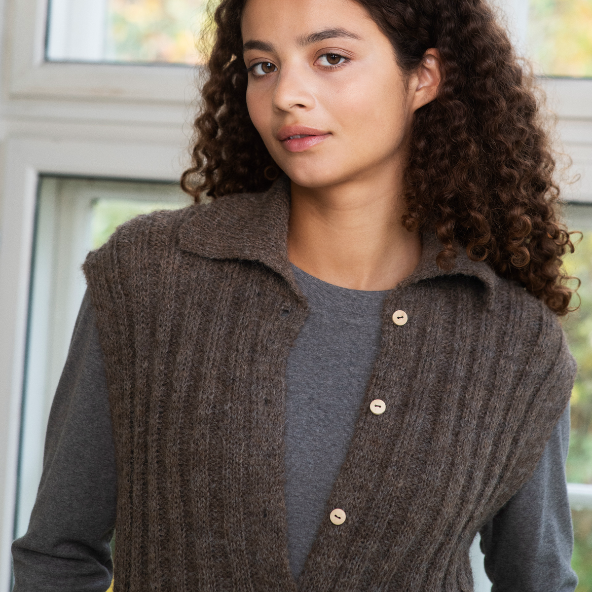 A woman wearing a brown ribbed knit button-front vest over a gray long-sleeve top, photographed indoors near a window.