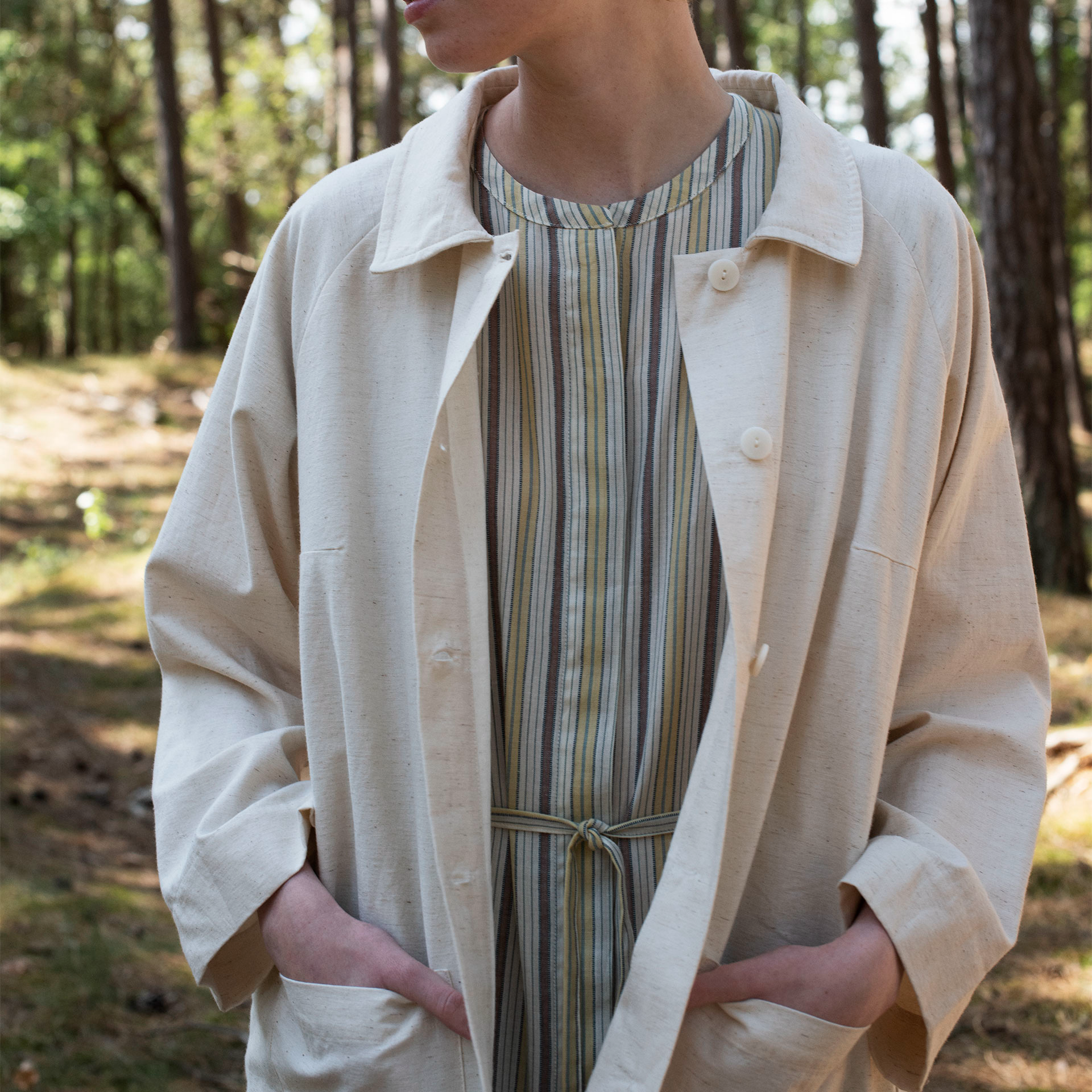 Person wearing an off white coat over a striped dress in a forest setting