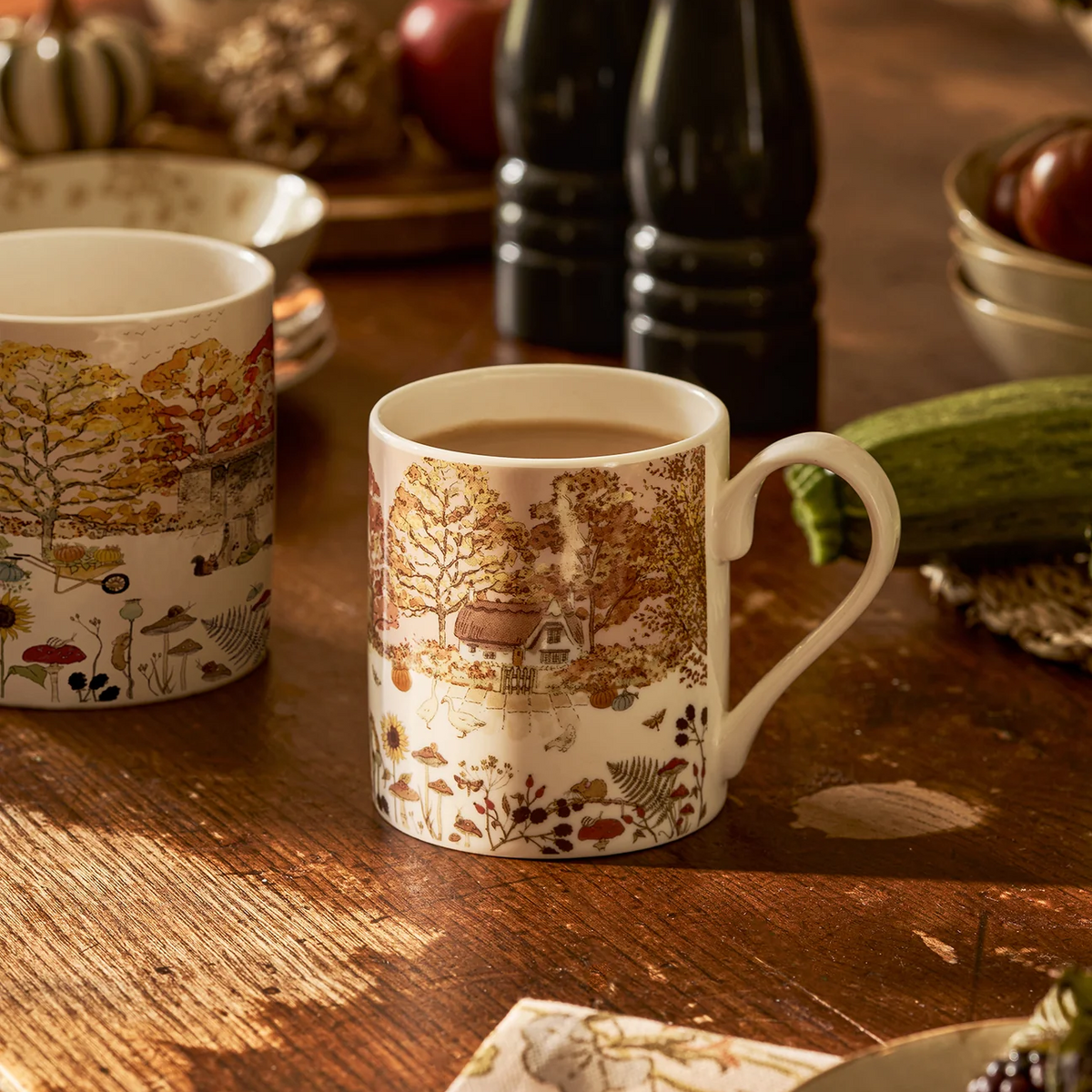 Two china mugs with an autumn forest and cottage landscape print, displayed on a rustic wooden table with seasonal decor in the background.