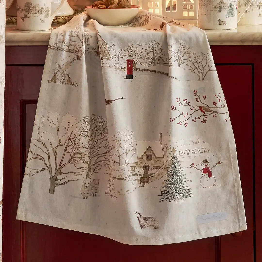 A kitchen tea towel with a winter village scene print, draped over a dark red cabinet. The print features snow-covered trees, a small house, a post box, a snowman, and a badger on an off-white background.