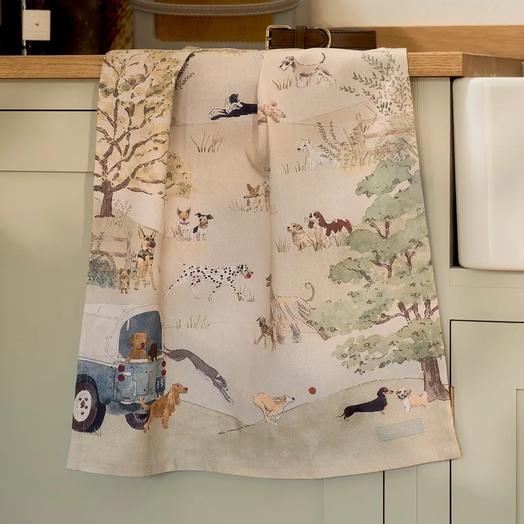 A cream-colored tea towel with a whimsical dog-themed illustration print, hanging over a light green kitchen cabinet.
