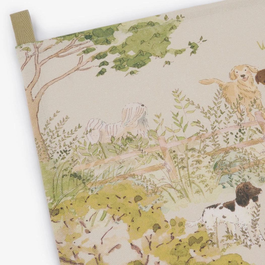 A close-up of a printed tea towel with a cream background and an illustrated outdoor scene featuring green foliage and dogs. A small green fabric tab is visible at the top left corner.