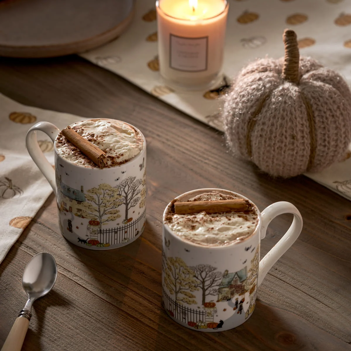 Two white china mugs decorated with a fall landscape illustration, filled with hot cocoa and topped with whipped cream and cinnamon sticks, set on a wooden table with a candle, a knitted pumpkin, and a spoon nearby.