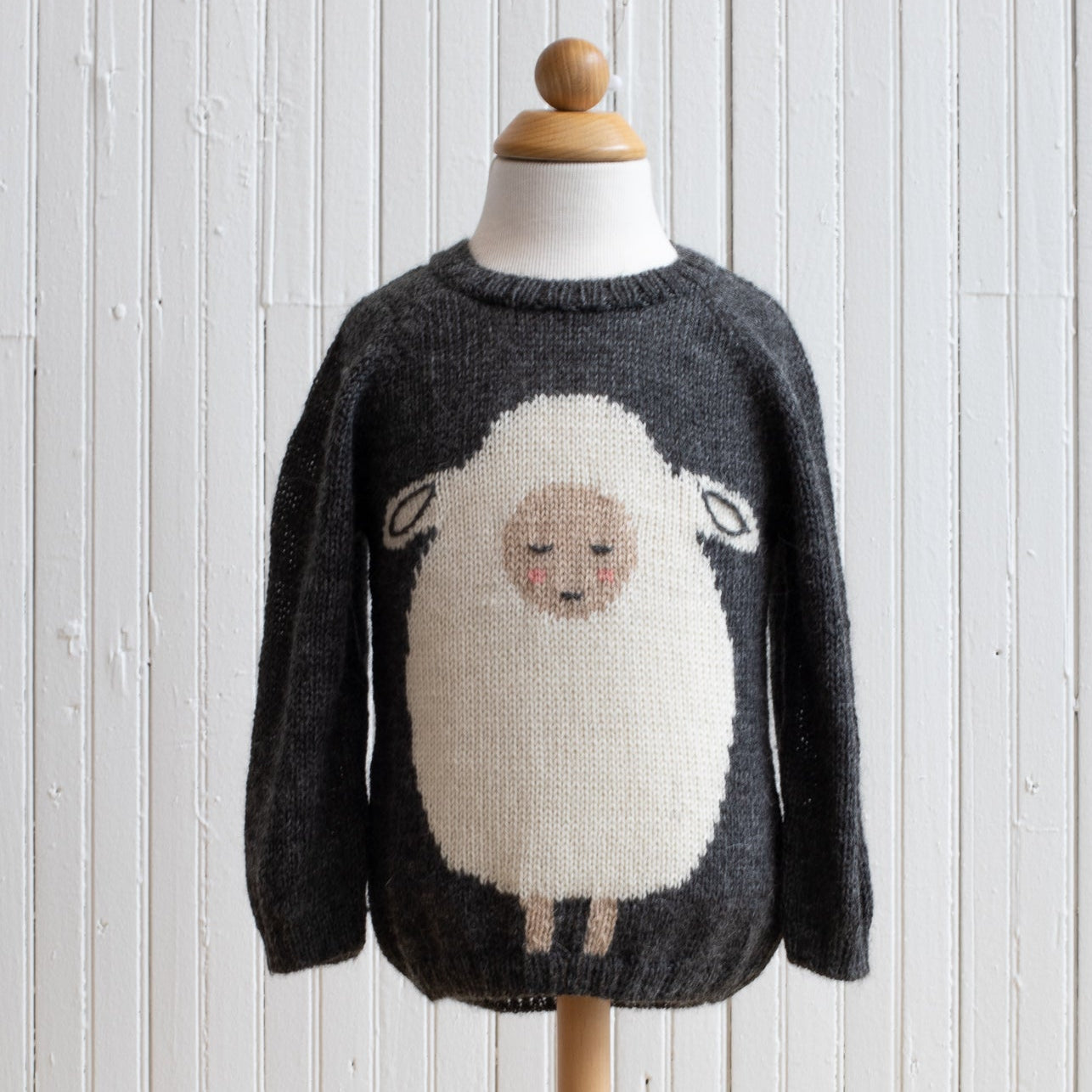 A gray baby alpaca sweater with a whimsical sheep design on the front, displayed on a wooden mannequin against a white wooden background.