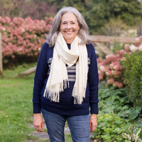 Woman standing in a garden wearing a navy sweater, striped shirt, and blue jeans with a white scarf.