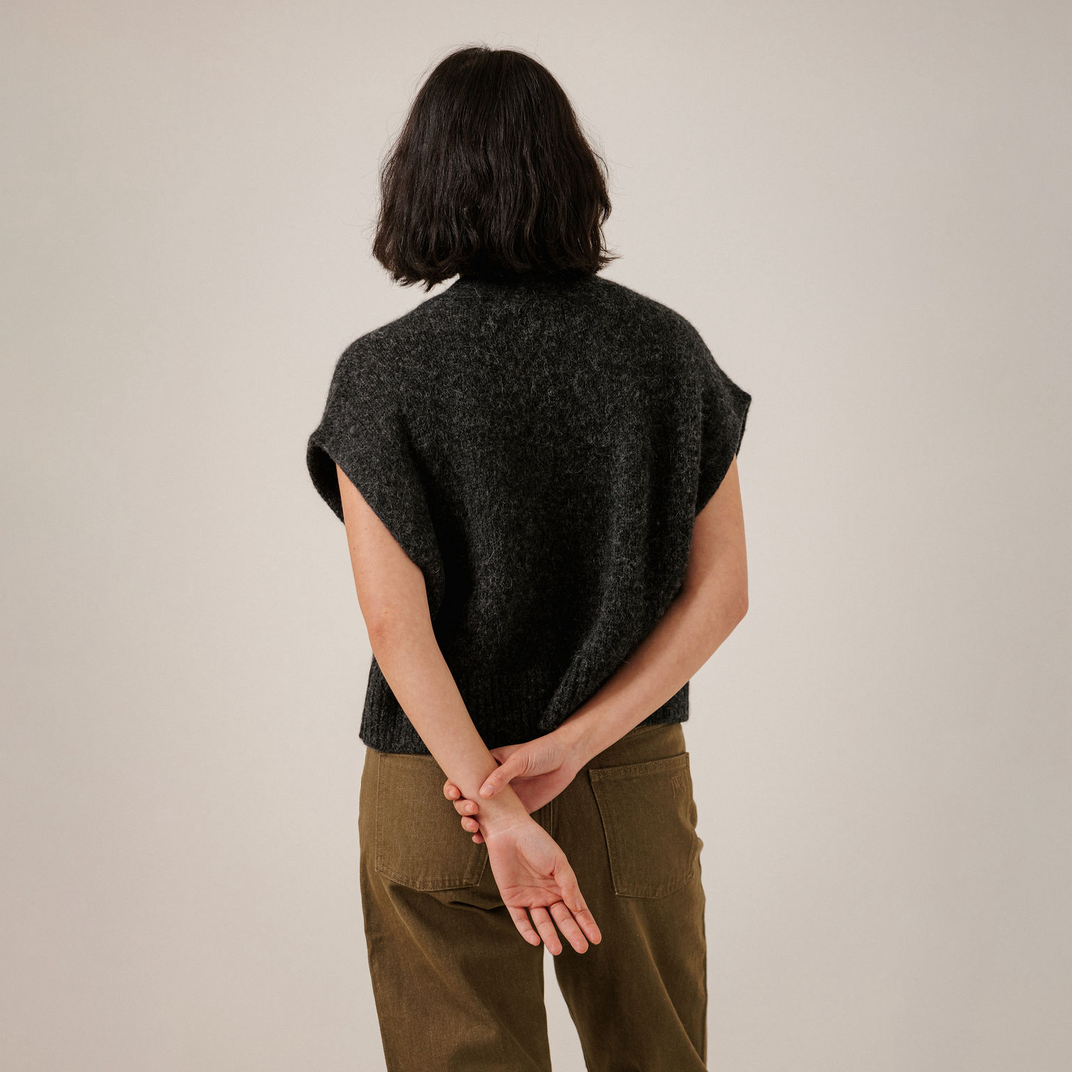 A person shown from the back wearing a dark charcoal heathered sleeveless knit top and olive green pants, standing against a plain light beige studio background.