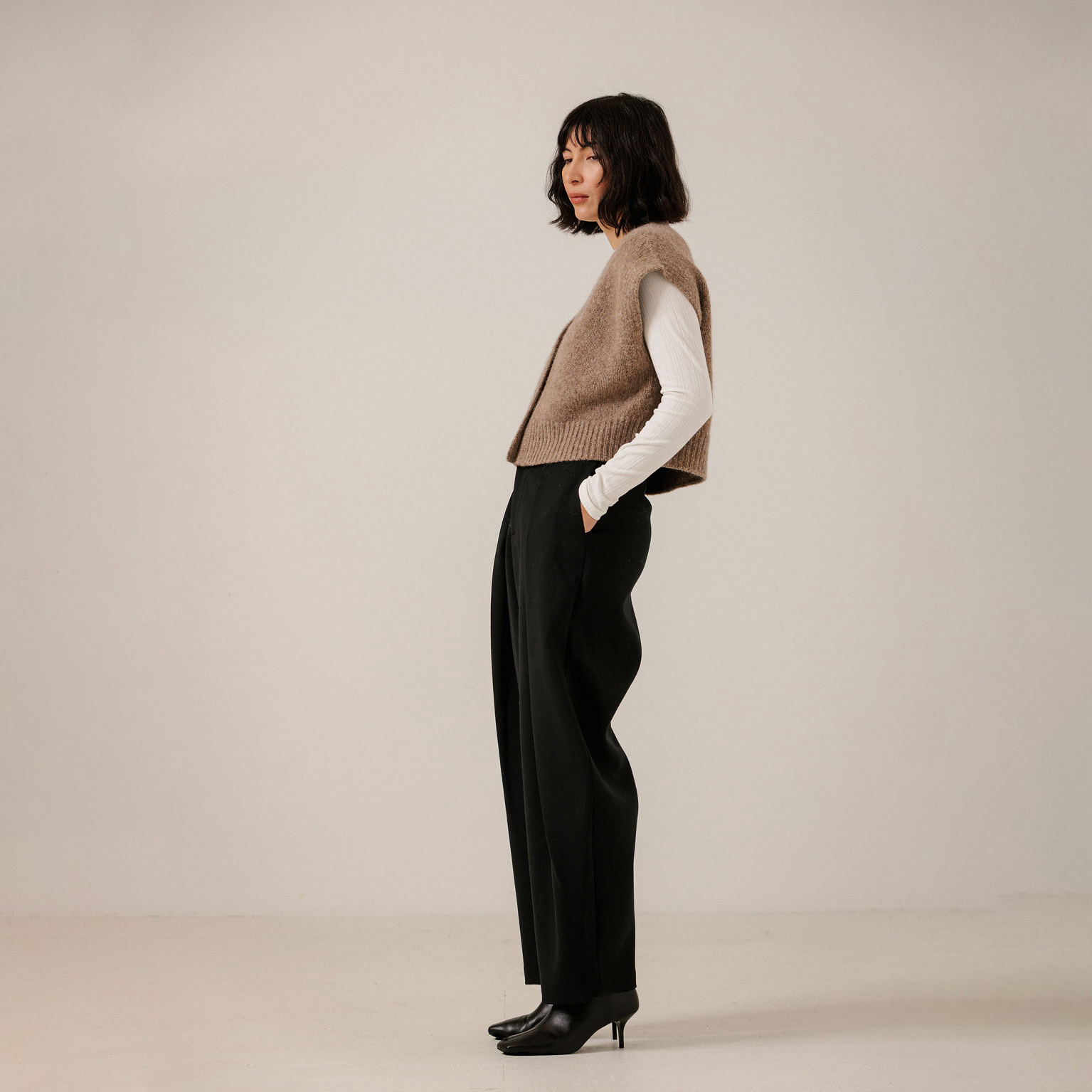 A woman stands in profile wearing a sleeveless taupe knit vest over a white long-sleeve top, black wide-leg trousers, and black pointed-toe heels against a plain light beige studio background.