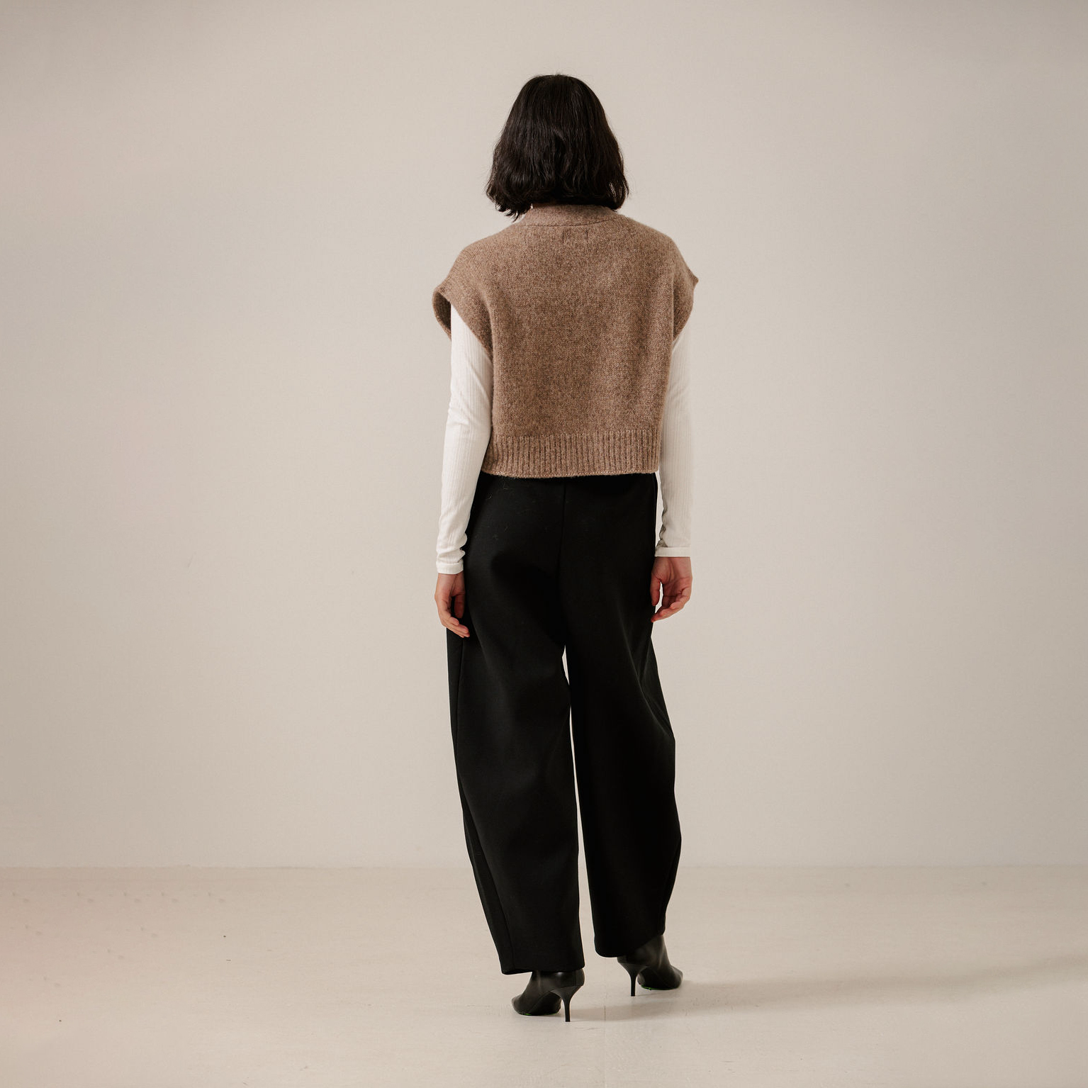 A model viewed from behind wearing a sleeveless taupe knit sweater vest over a white long-sleeve shirt and black wide-leg trousers, standing against a neutral off-white studio background.