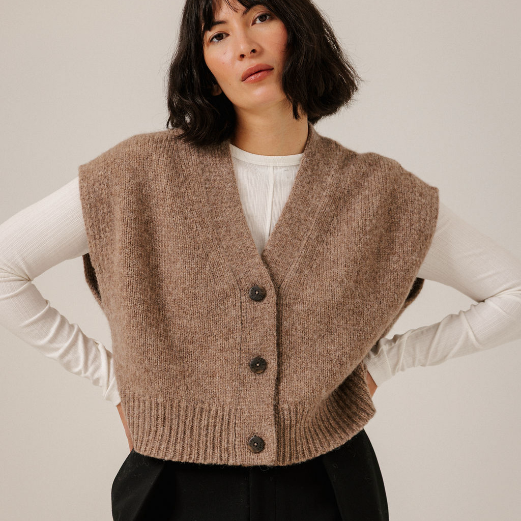 A woman wearing a brown ribbed knit cardigan vest over a white long-sleeve top and black pants, photographed against a plain light beige studio background.