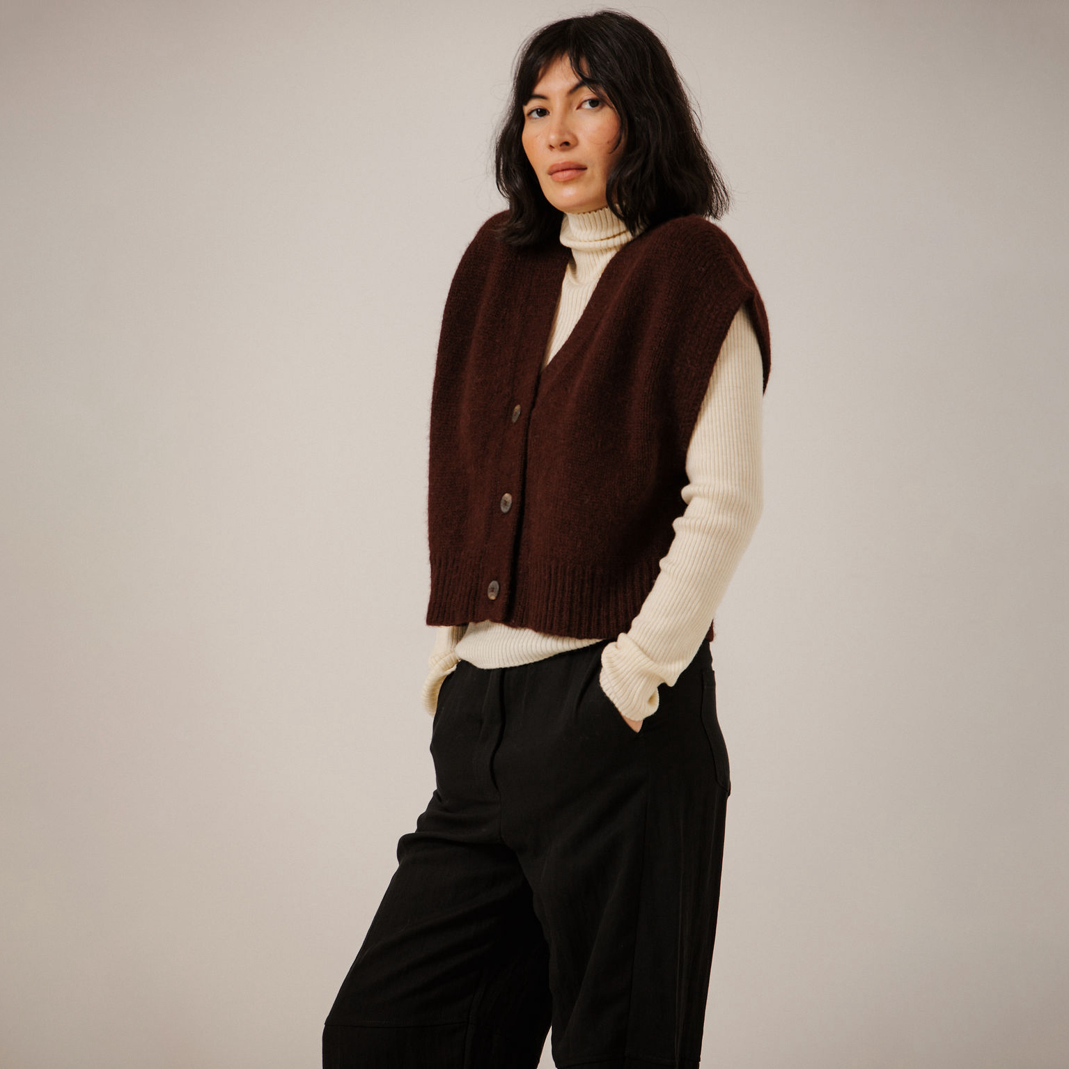 A woman wearing a dark maroon ribbed knit vest over a cream turtleneck sweater and black trousers, standing against a plain light gray studio background.