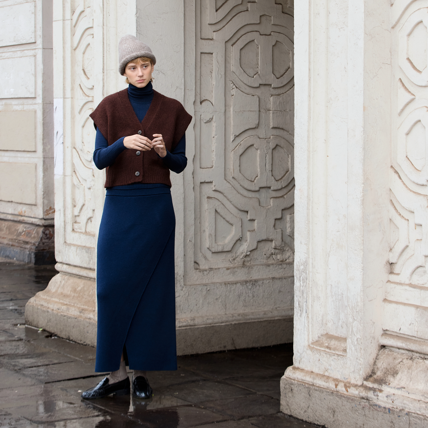 A model stands in a doorway wearing a dark maroon sleeveless knit vest over a navy blue turtleneck, paired with a long navy wrap skirt and black shoes.