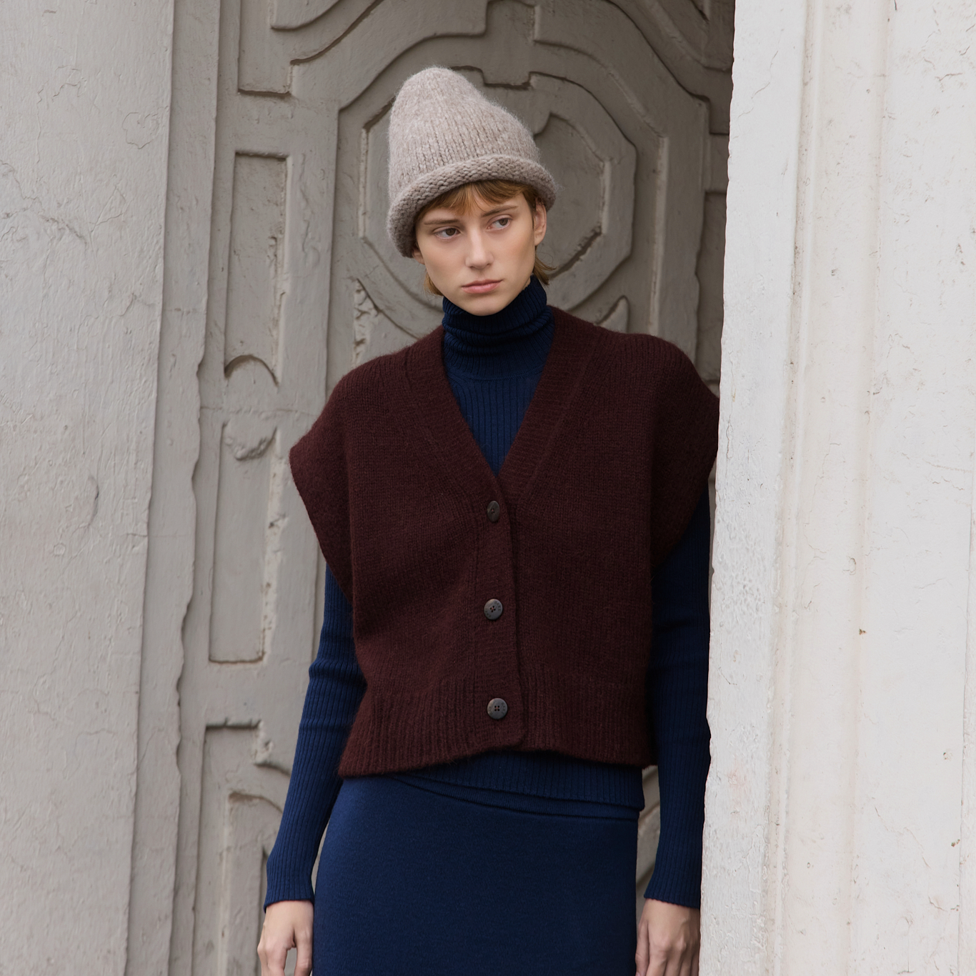 A model wearing a dark maroon sleeveless button-front knit vest over a navy blue turtleneck and matching skirt, paired with a beige knit beanie, standing in a doorway.
