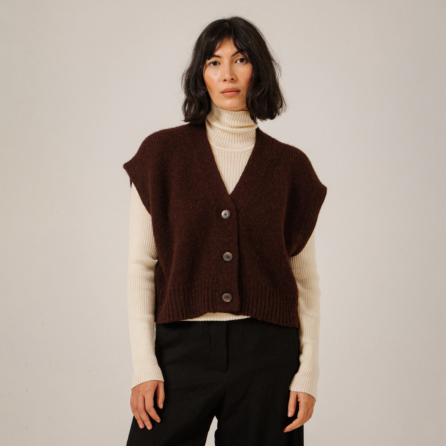 A model wearing a dark maroon sleeveless knit cardigan vest over a cream ribbed turtleneck sweater and black trousers, standing against a neutral light gray background.