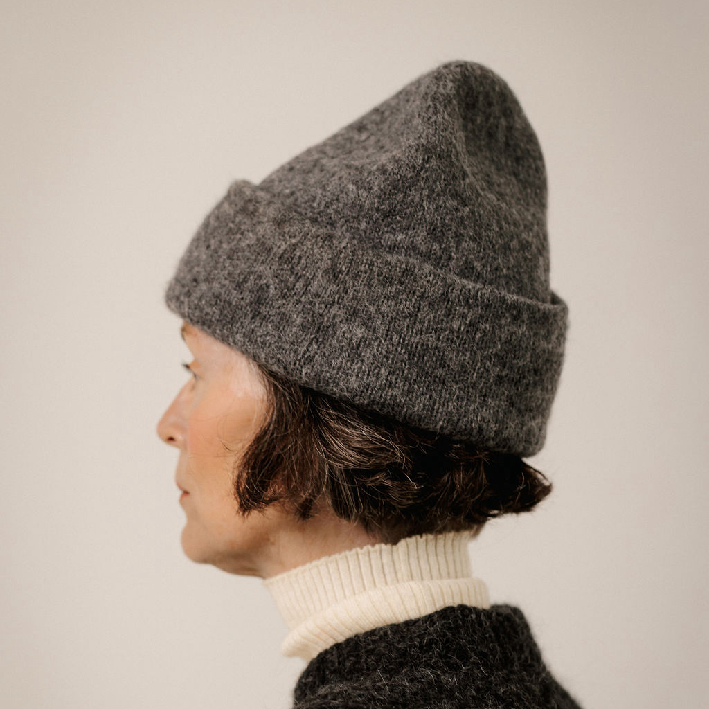 A person shown in side profile wearing a dark heather gray knit beanie and a cream ribbed turtleneck sweater against a plain light beige background.