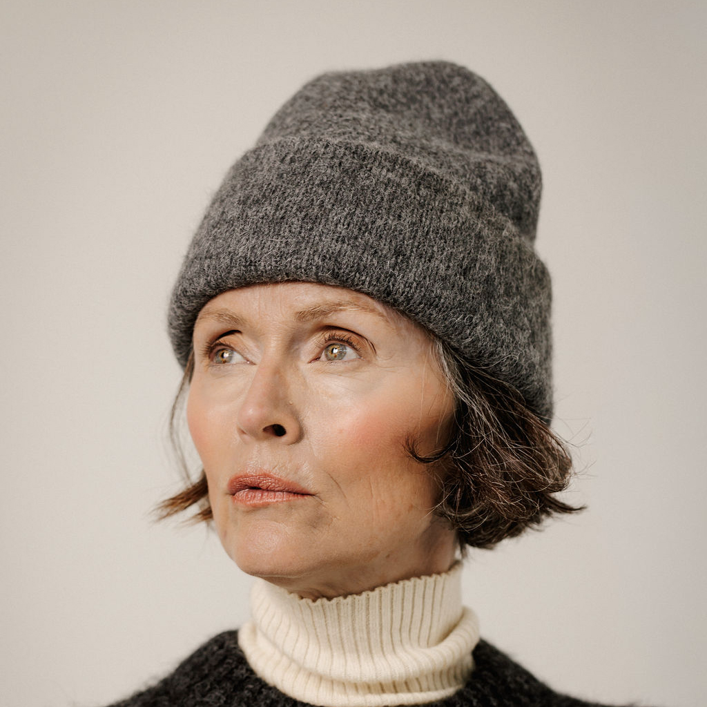 A close-up portrait of a woman wearing a dark gray heathered knit beanie and a cream ribbed turtleneck sweater against a plain light background.