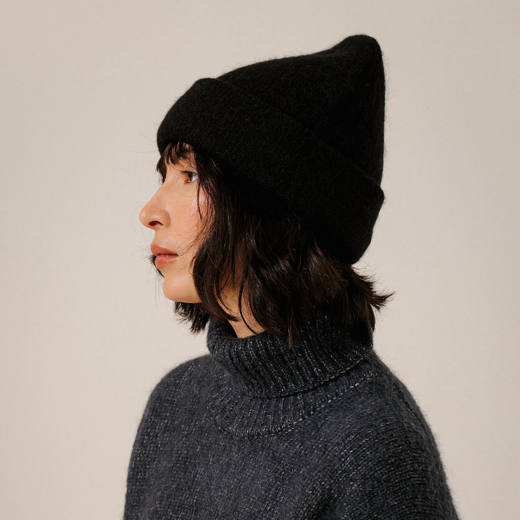 A person shown in side profile wearing a black knit beanie and a dark charcoal turtleneck sweater against a plain light beige background.