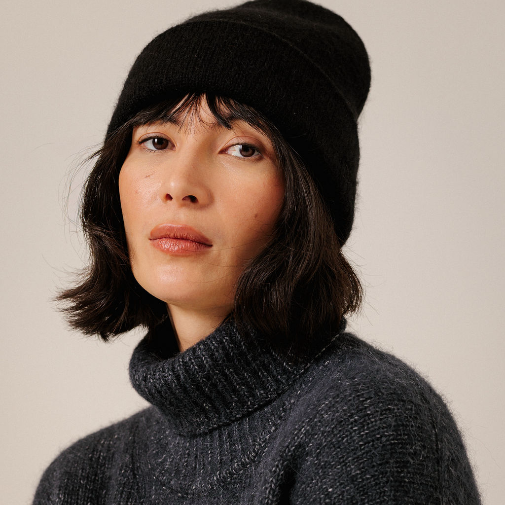A close-up portrait of a woman wearing a black knit beanie and a dark charcoal gray turtleneck sweater against a plain light beige background.