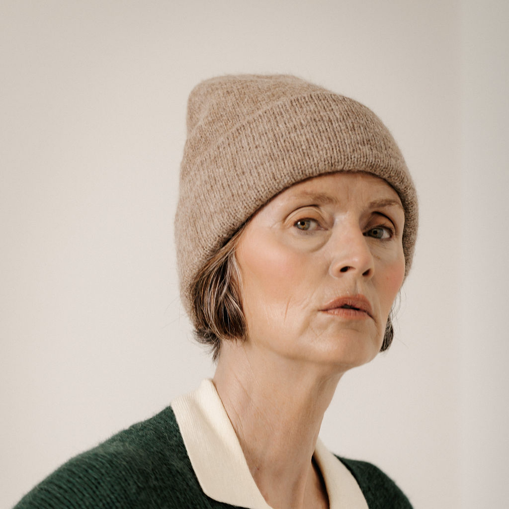 A close-up portrait of a woman wearing a beige knit beanie and a dark green sweater with a cream collar, set against a plain light background.