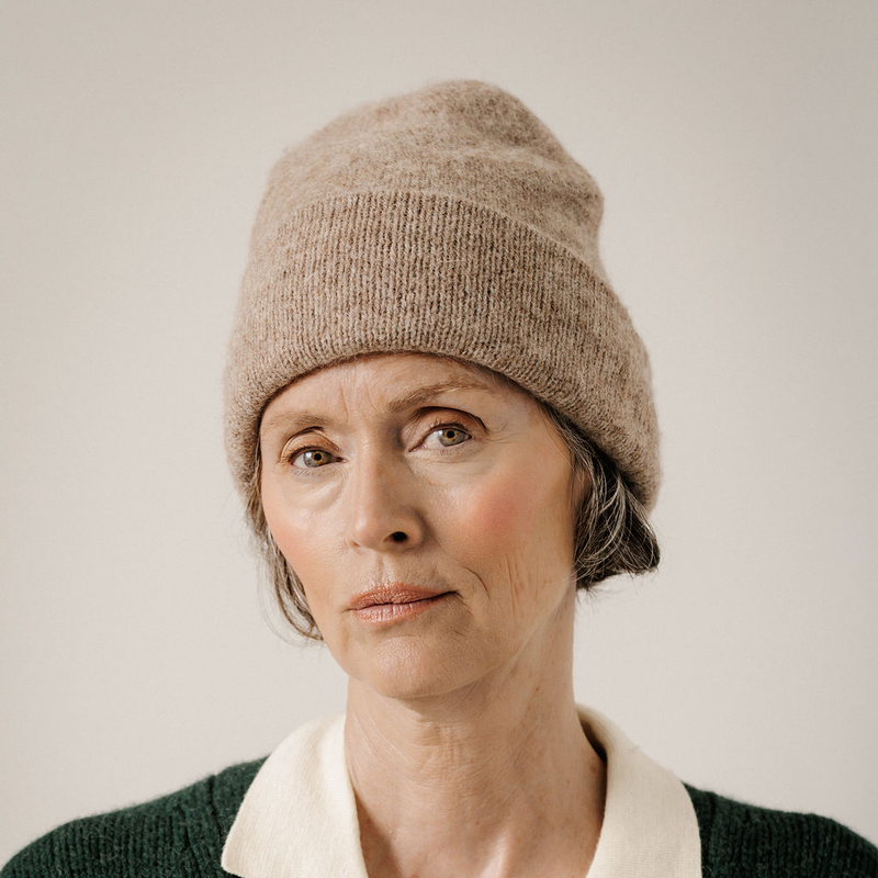 A close-up portrait of a woman wearing a beige knit beanie and a dark green sweater with a cream collar, set against a plain light background.