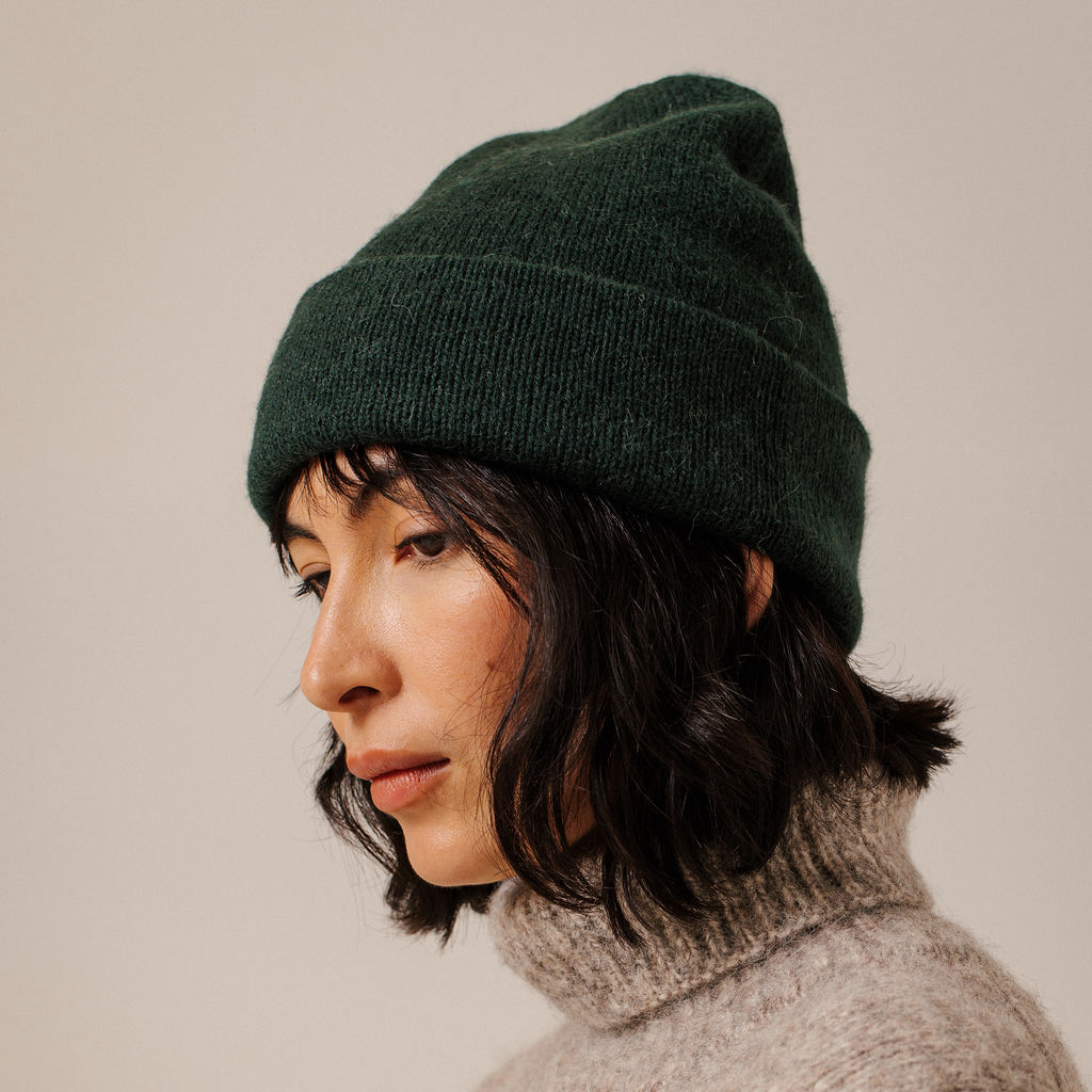 A person shown in profile wearing a dark forest green knit beanie and a light gray turtleneck sweater against a plain beige studio background.