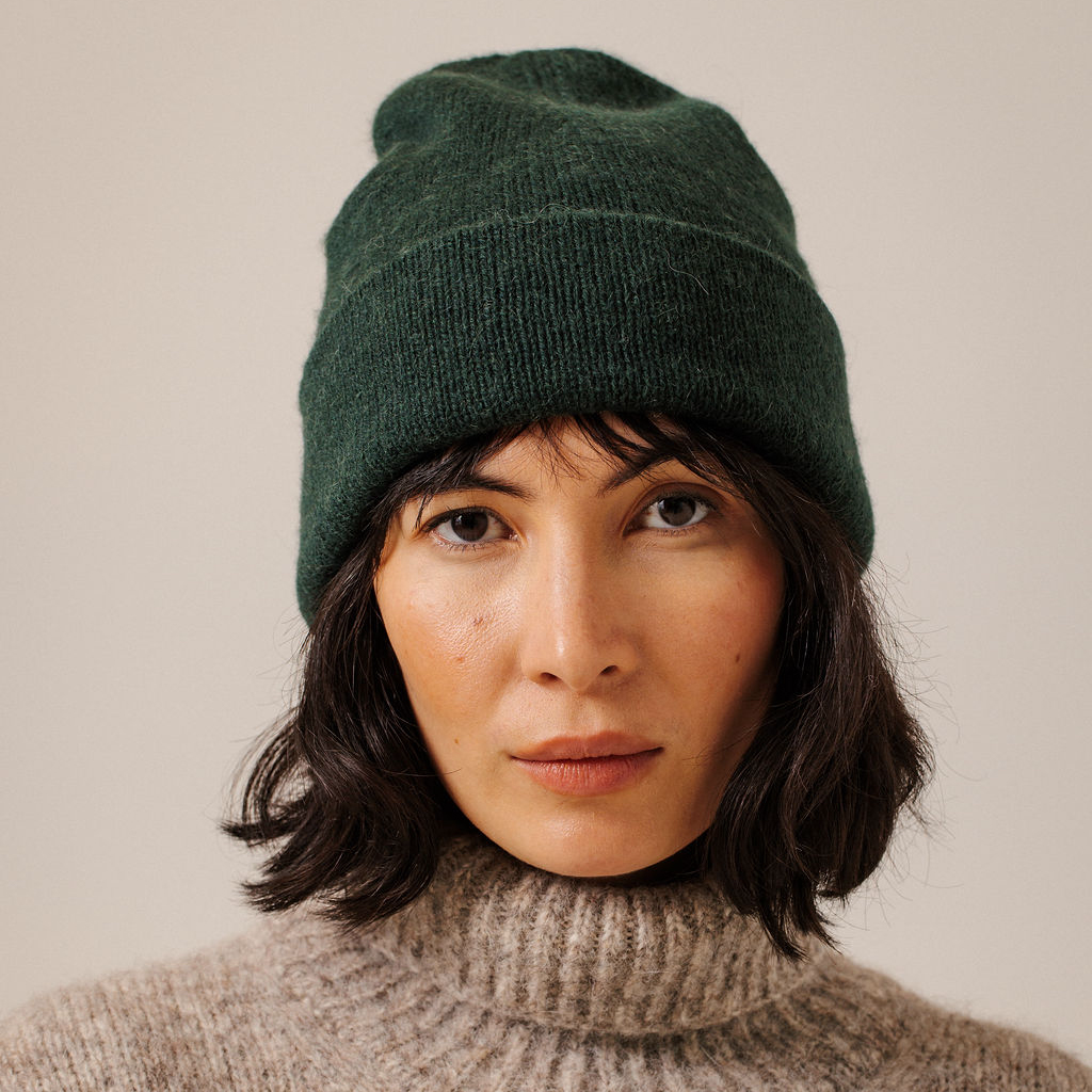 A woman wearing a dark forest green knit beanie and a beige ribbed turtleneck sweater, photographed against a plain light beige background.