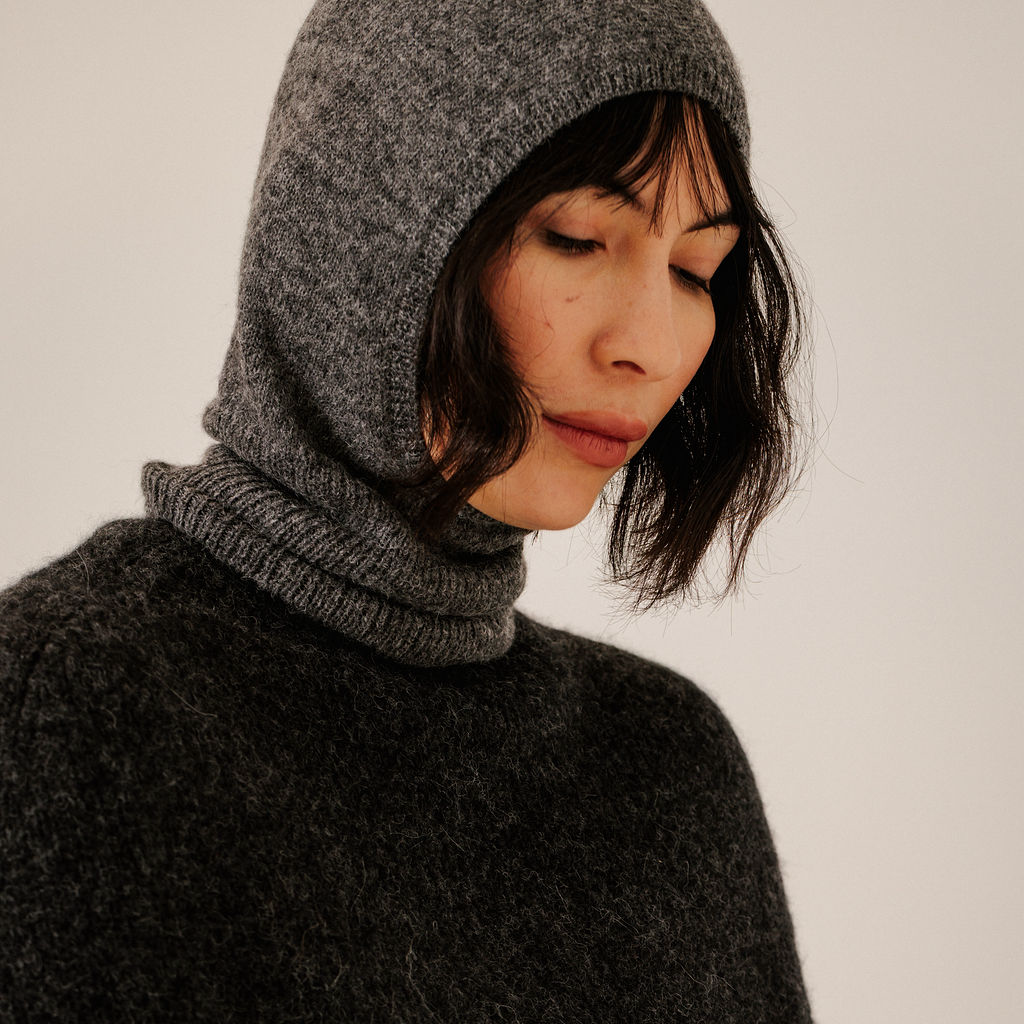 A close-up of a woman wearing a dark gray heathered knit hooded balaclava and a matching dark gray fuzzy sweater against a plain light background.