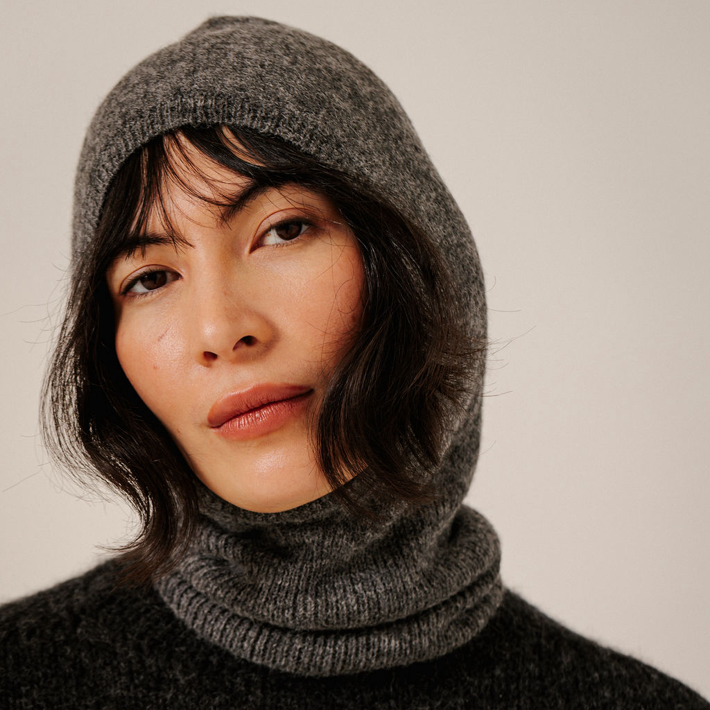 A close-up of a woman wearing a dark gray heathered knit hooded balaclava and a matching dark gray fuzzy sweater against a plain light background.