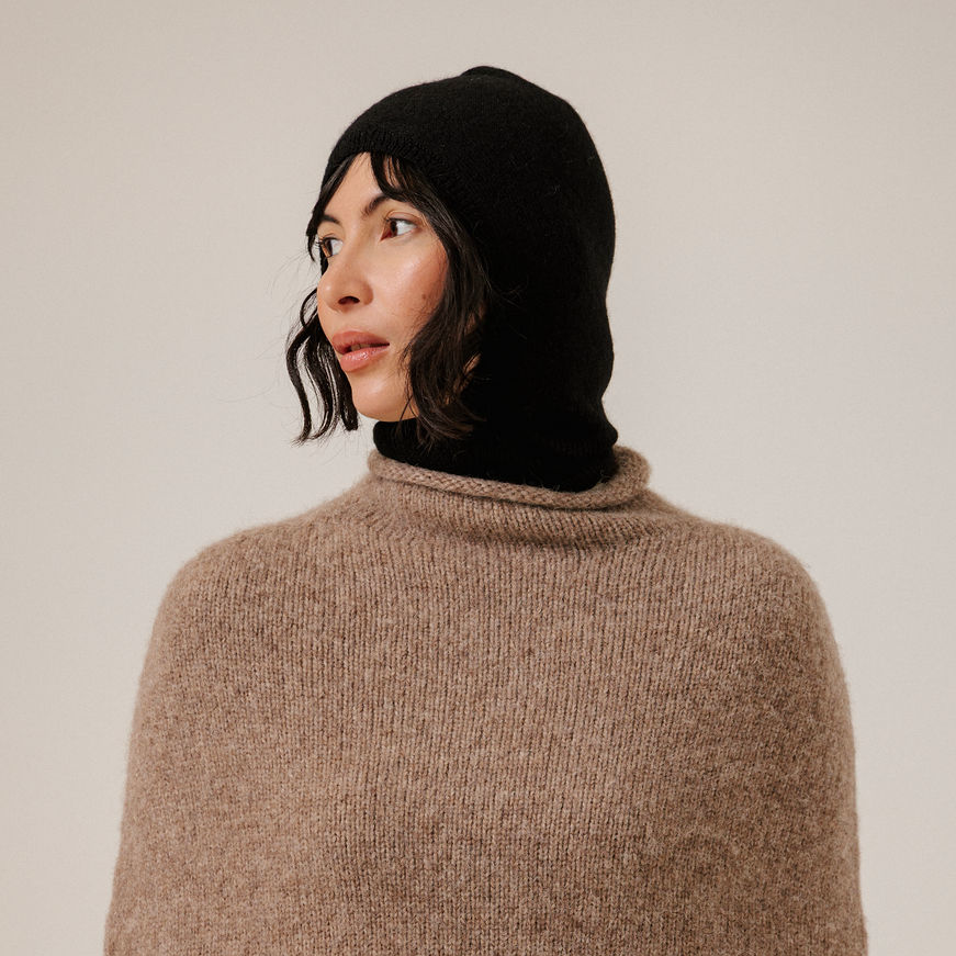 A close-up of a woman wearing a black knit hooded balaclava and a beige knit poncho against a plain light background.