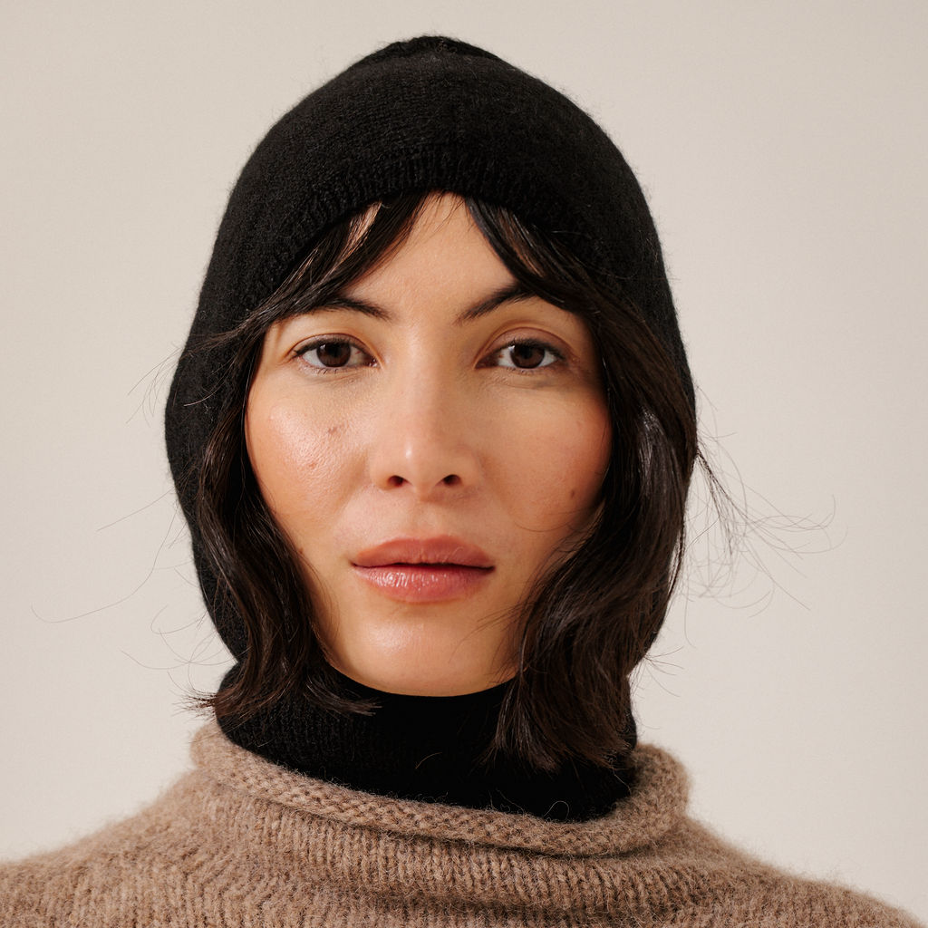 A close-up of a woman wearing a black knit hooded balaclava and a beige knit top against a plain light background.