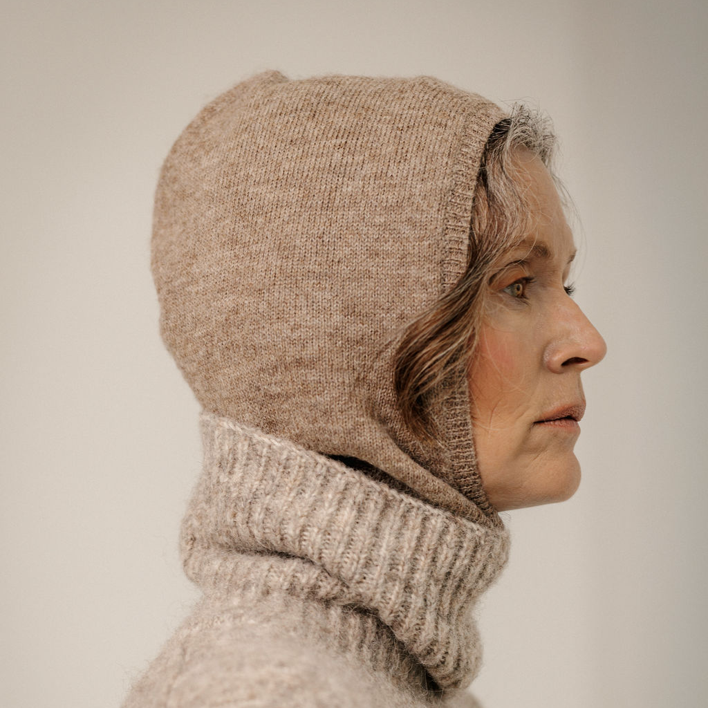 A close-up portrait of a woman wearing a beige knit balaclava and matching ribbed turtleneck sweater against a soft light gray background.