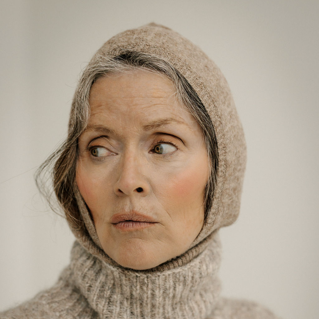 A close-up portrait of a woman wearing a beige knit balaclava and matching ribbed turtleneck sweater against a soft light gray background.