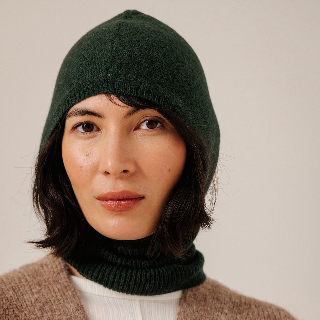 A close-up of a woman wearing a forest green knit hooded balaclava and a beige knit top against a plain light background.