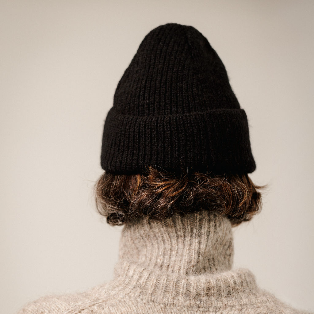 A person shown from the back wearing a black ribbed knit beanie, a beige turtleneck sweater, and short brown hair, against a plain light beige background.