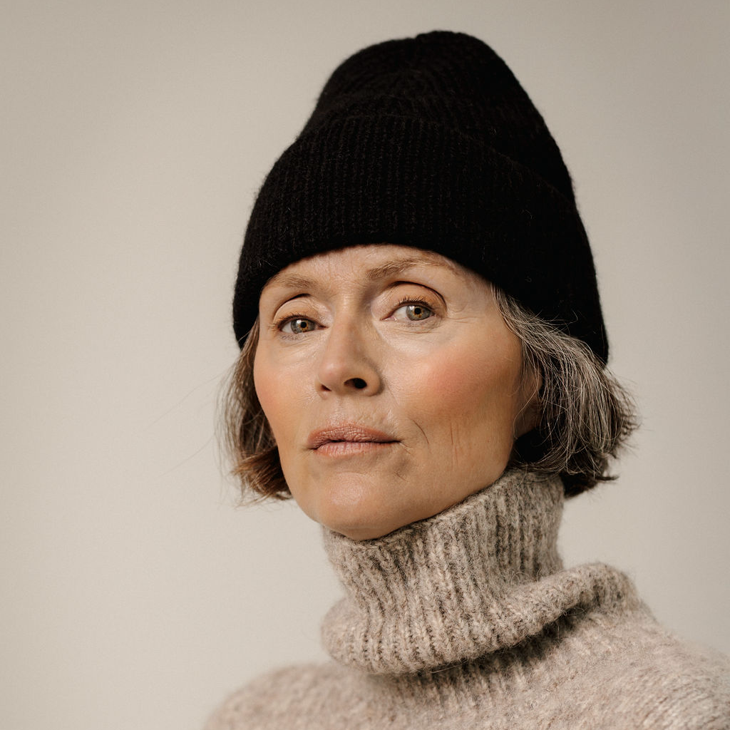A close-up portrait of a woman wearing a black ribbed knit beanie and a beige turtleneck sweater against a plain light gray background.
