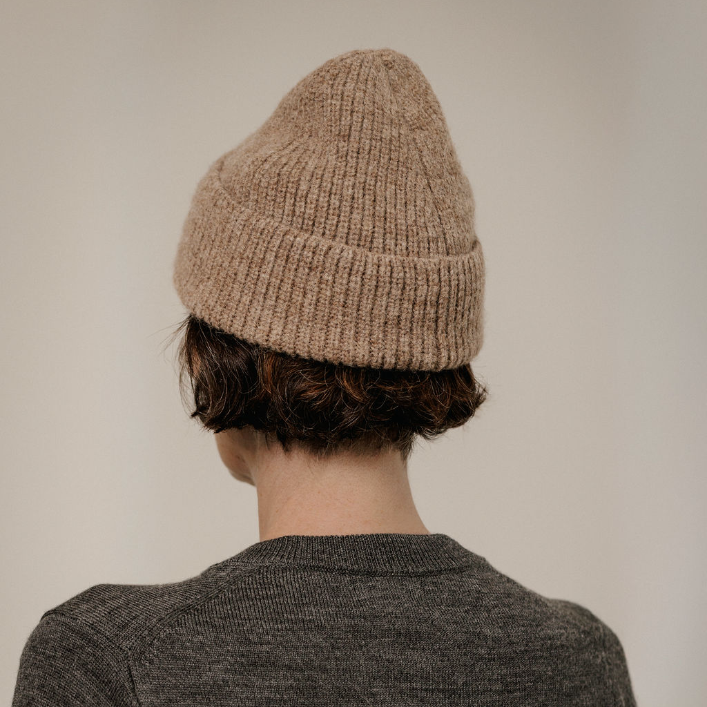 A person shown from the back wearing a beige ribbed knit beanie and a dark gray knit sweater against a plain light gray background.