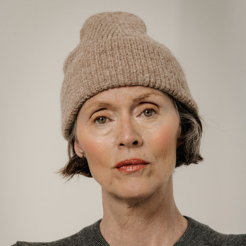 A close-up portrait of a woman wearing a chunky knit beige beanie hat against a plain light gray background.
