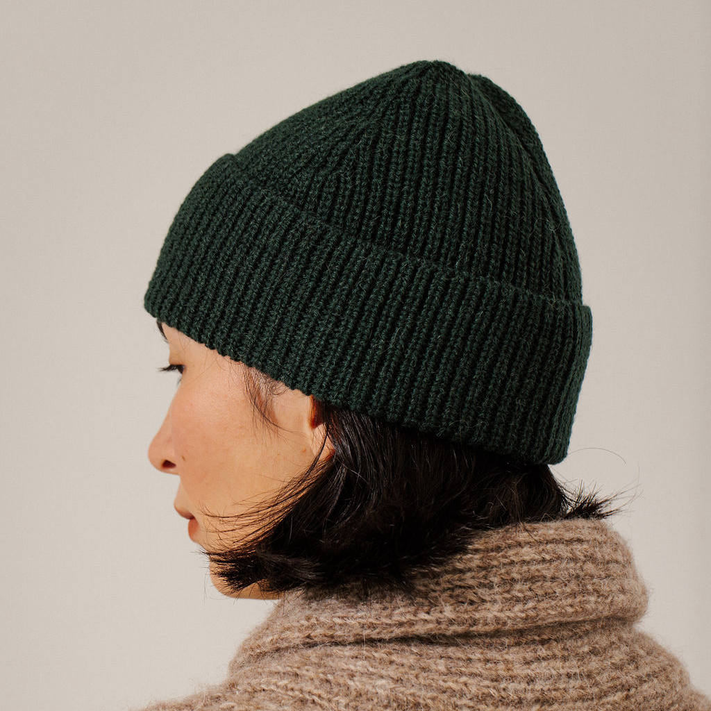 A woman wearing a dark forest green ribbed knit beanie and a chunky taupe knit sweater over a cream ribbed turtleneck, photographed against a neutral light gray background.
