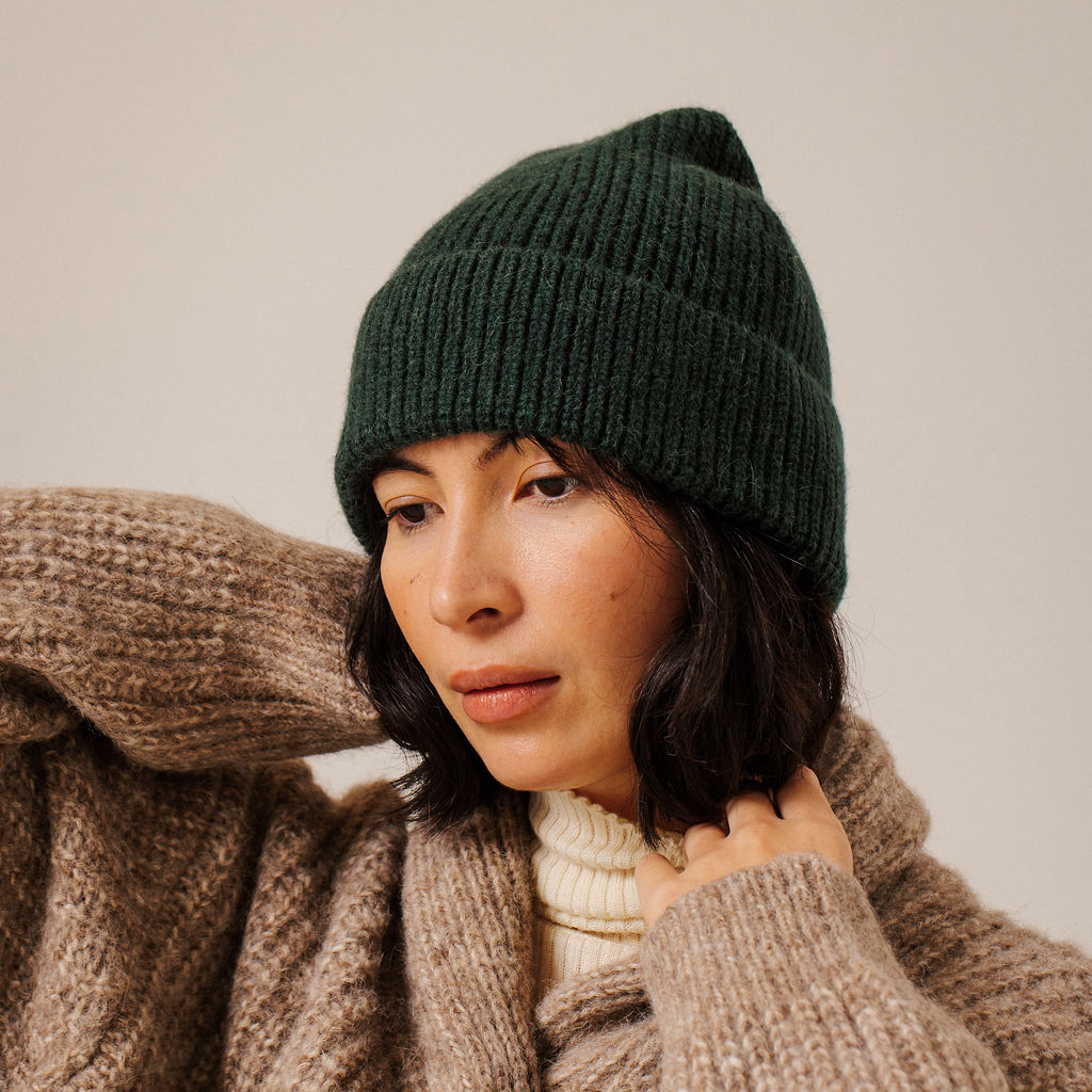 A woman wearing a dark forest green ribbed knit beanie hat and a chunky beige knit sweater, photographed against a plain light beige background.