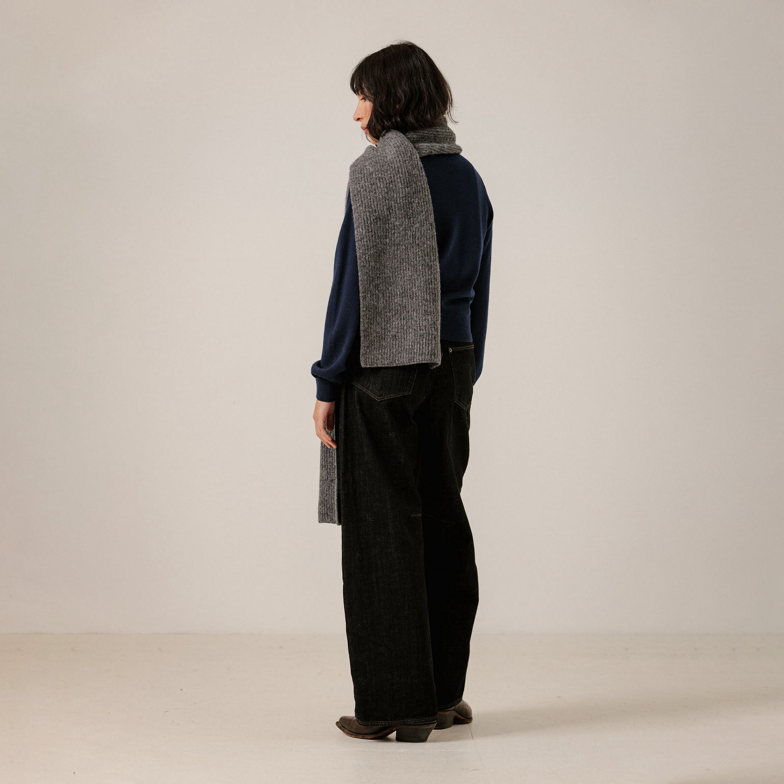 A model is shown from the back wearing a navy blue long-sleeve top, black wide-leg trousers, and a gray textured knit scarf draped over their shoulders, set against a plain off-white studio background.